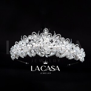 Puede incluir: Una tiara plateada con cristales transparentes y perlas blancas. La corona presenta un diseño floral con un pico central y una banda curva. El texto "LA CASA JEWELRY" se muestra debajo.