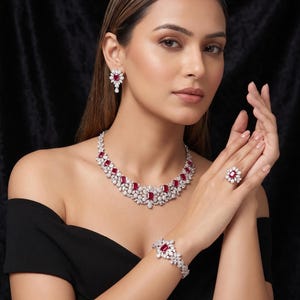Puede incluir: Un conjunto de joyas completo que incluye collar, pendientes, pulsera y anillo. El conjunto presenta piedras claras y brillantes y gemas rojas vibrantes. Las joyas se exhiben sobre un fondo negro, resaltando el intrincado diseño y el brillo.