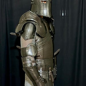 May include: A full suit of dark metal armor, including a helmet with a cross design, shoulder guards, and arm and leg protection. The armor is riveted and features leather straps and chainmail accents. The overall design is medieval.