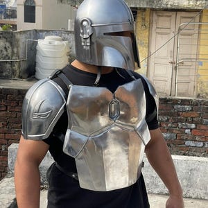 May include: A silver-colored Mandalorian helmet and chest armor set. The helmet has a T-shaped visor and ear pieces. The chest armor is made of multiple metal plates and is secured with black straps. The armor is worn over a black shirt.
