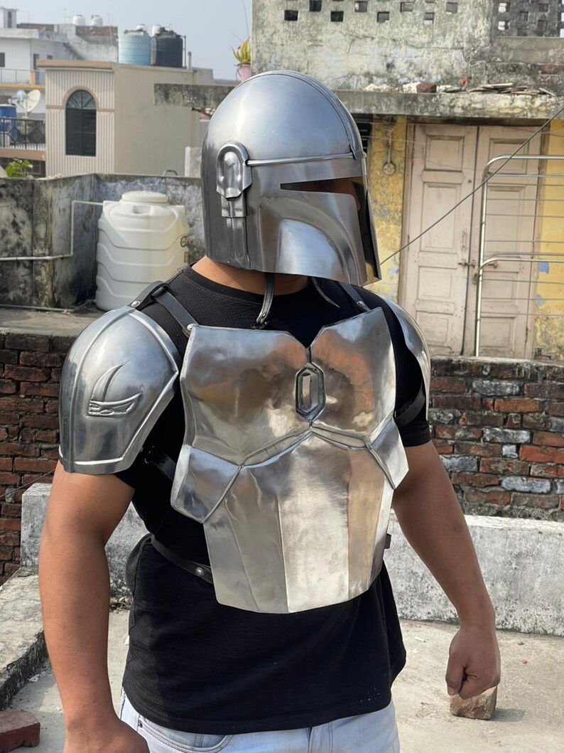 May include: A silver-colored Mandalorian helmet and chest armor set. The helmet has a T-shaped visor and ear pieces. The chest armor is made of multiple metal plates and is secured with black straps. The armor is worn over a black shirt.