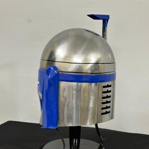 Op de afbeelding: Een zilverkleurige helm met blauwe accenten, geplaatst op een zwarte standaard. De helm heeft een afgeronde bovenkant en verticale lijnen aan de zijkant. Het ontwerp suggereert een futuristisch thema.