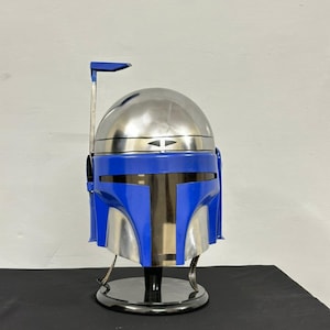 Op de afbeelding: Een replica van een Mandalorian-helm in blauw en zilver, tentoongesteld op een zwarte standaard. De helm heeft een afgeronde bovenkant, een T-vormig vizier en een afstandsmeter. Het kleurenschema is een mix van metallic zilver en levendig blauw.