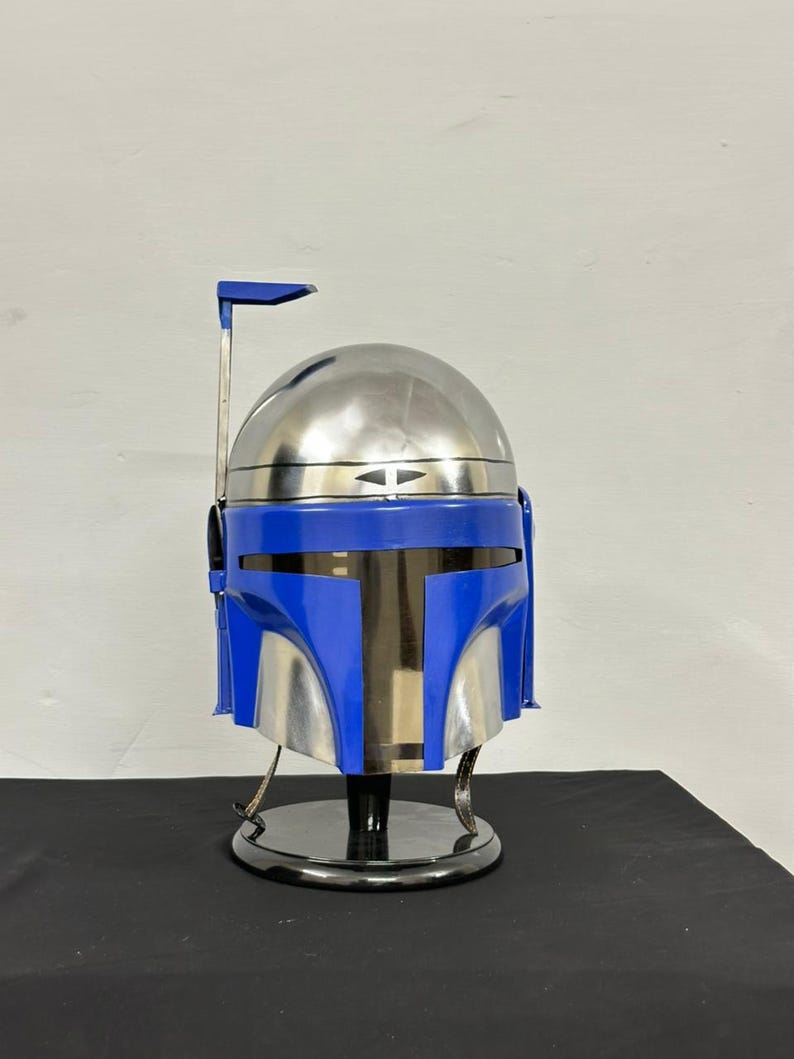 Op de afbeelding: Een replica van een Mandalorian-helm in blauw en zilver, tentoongesteld op een zwarte standaard. De helm heeft een afgeronde bovenkant, een T-vormig vizier en een afstandsmeter. Het kleurenschema is een mix van metallic zilver en levendig blauw.