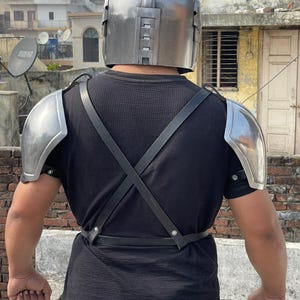 May include: A silver-colored helmet and shoulder armor set. The helmet has a rounded top with a series of rectangular details on the sides. The shoulder armor is curved and metallic. Black straps cross the back and secure the armor.