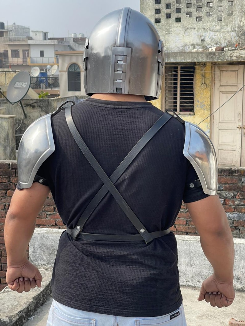 May include: A silver-colored helmet and shoulder armor set. The helmet has a rounded top with a series of rectangular details on the sides. The shoulder armor is curved and metallic. Black straps cross the back and secure the armor.