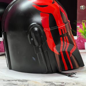 May include: A black helmet with a red graphic design on the front and top. The helmet has a visor and is likely made of plastic or a similar material. The design is symmetrical and appears to be painted on.