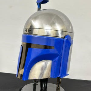 Op de afbeelding: Een zilveren en blauwe helm, waarschijnlijk een rekwisiet of verzamelobject, wordt tentoongesteld op een zwarte standaard. De helm heeft een geborstelde metalen afwerking op het bovenste gedeelte en een felblauwe voorkant. Een kleine antenne steekt uit de bovenkant.