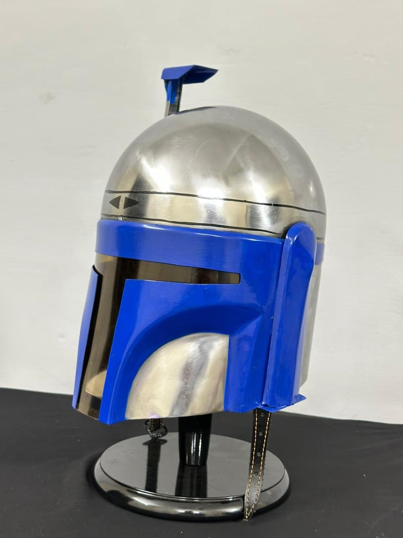 Op de afbeelding: Een zilveren en blauwe helm, waarschijnlijk een rekwisiet of verzamelobject, wordt tentoongesteld op een zwarte standaard. De helm heeft een geborstelde metalen afwerking op het bovenste gedeelte en een felblauwe voorkant. Een kleine antenne steekt uit de bovenkant.