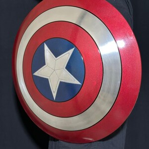 May include: A red, white, and blue Captain America shield. The shield has a metallic silver ring around the perimeter and a white star in the center of a blue circle. The shield is round and has a glossy finish.