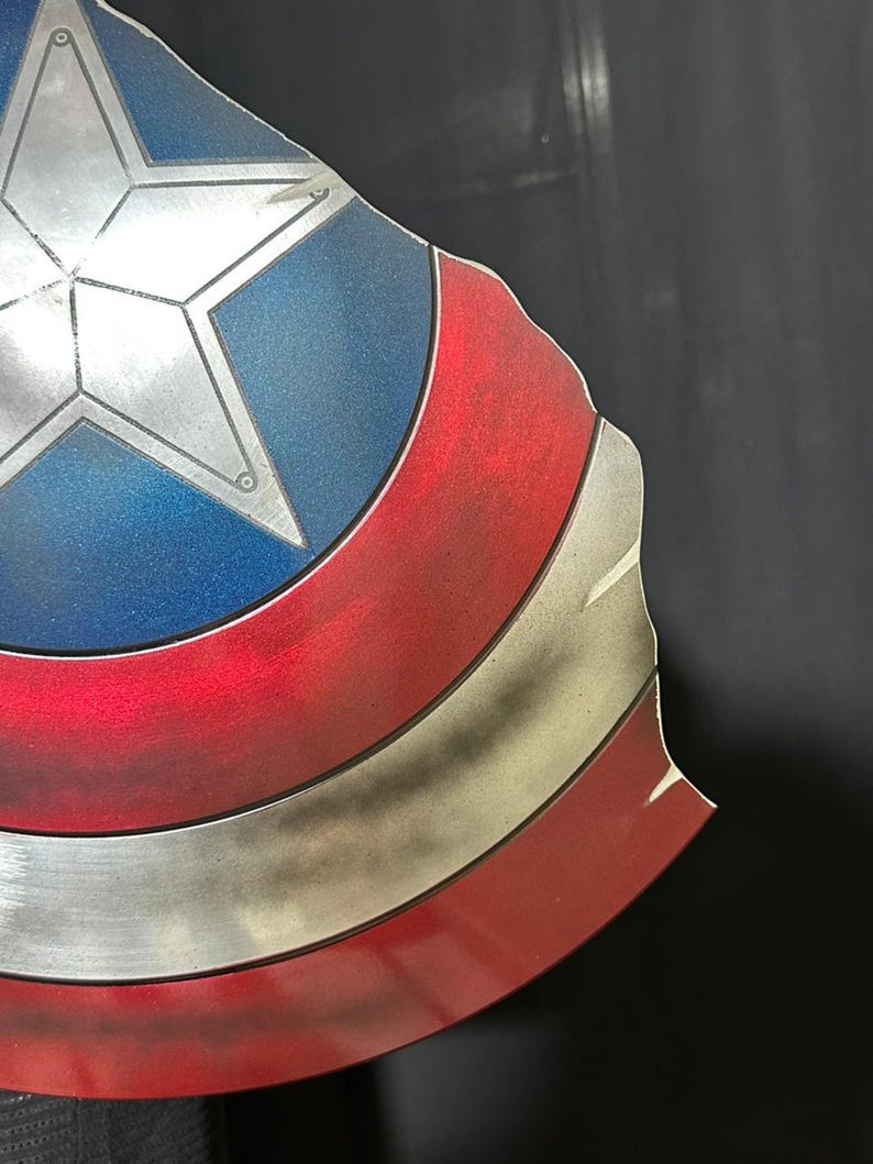 Może przedstawiać: Zbliżenie tarczy Kapitana Ameryki. Tarcza posiada srebrną gwiazdę na niebieskim tle, z czerwonymi i białymi paskami. Tarcza ma metaliczne wykończenie i lekko postarzany wygląd.