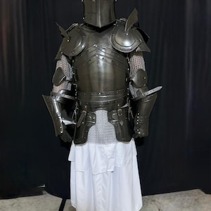 May include: Full suit of dark metal armor, including a helmet, chest plate, shoulder guards, and arm protection. The armor is worn over chainmail and a white robe. The helmet has a cross design. The person is wearing white sandals.