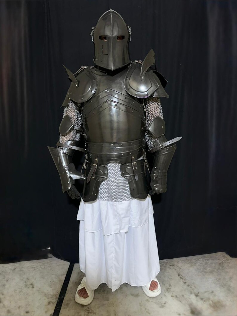 May include: Full suit of dark metal armor, including a helmet, chest plate, shoulder guards, and arm protection. The armor is worn over chainmail and a white robe. The helmet has a cross design. The person is wearing white sandals.