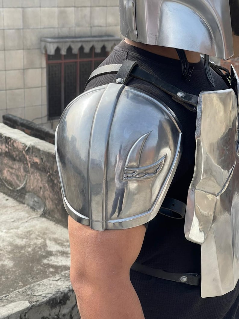 May include: Silver-colored armor set, including a helmet and shoulder pads, with a stylized emblem. The armor is worn over a black shirt. The shoulder pads are attached with black straps.