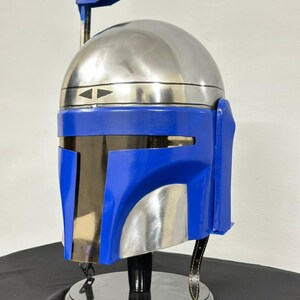 Op de afbeelding: Een helm in Mandalorian-stijl met een gepolijste zilveren koepel en een felblauwe frontplaat. De helm heeft een T-vormig vizier, een zwarte rand en een zwarte standaard. Het ontwerp omvat een afstandsmeter en een riem.