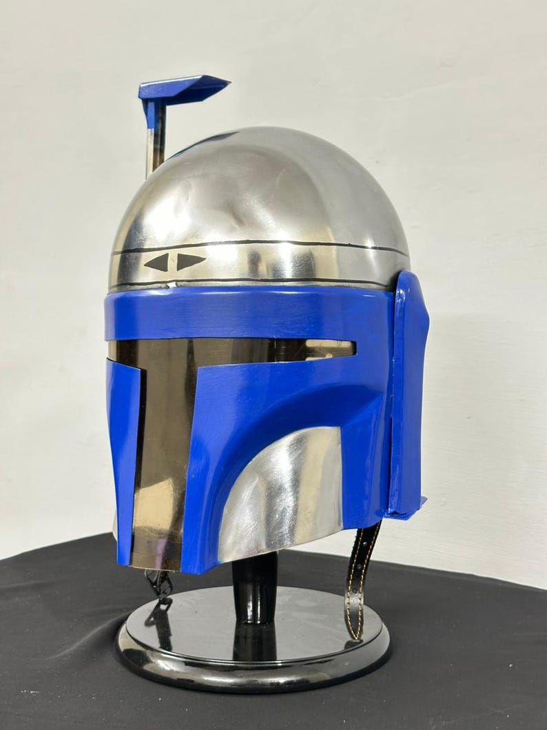 Op de afbeelding: Een helm in Mandalorian-stijl met een gepolijste zilveren koepel en een felblauwe frontplaat. De helm heeft een T-vormig vizier, een zwarte rand en een zwarte standaard. Het ontwerp omvat een afstandsmeter en een riem.