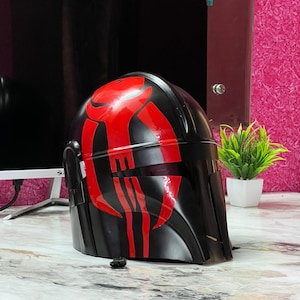 May include: A black helmet with red markings, likely a costume piece. The helmet has a smooth, glossy finish and a visor. The red design is a stylized pattern on the front and top of the helmet. The helmet is sitting on a marble-patterned surface.