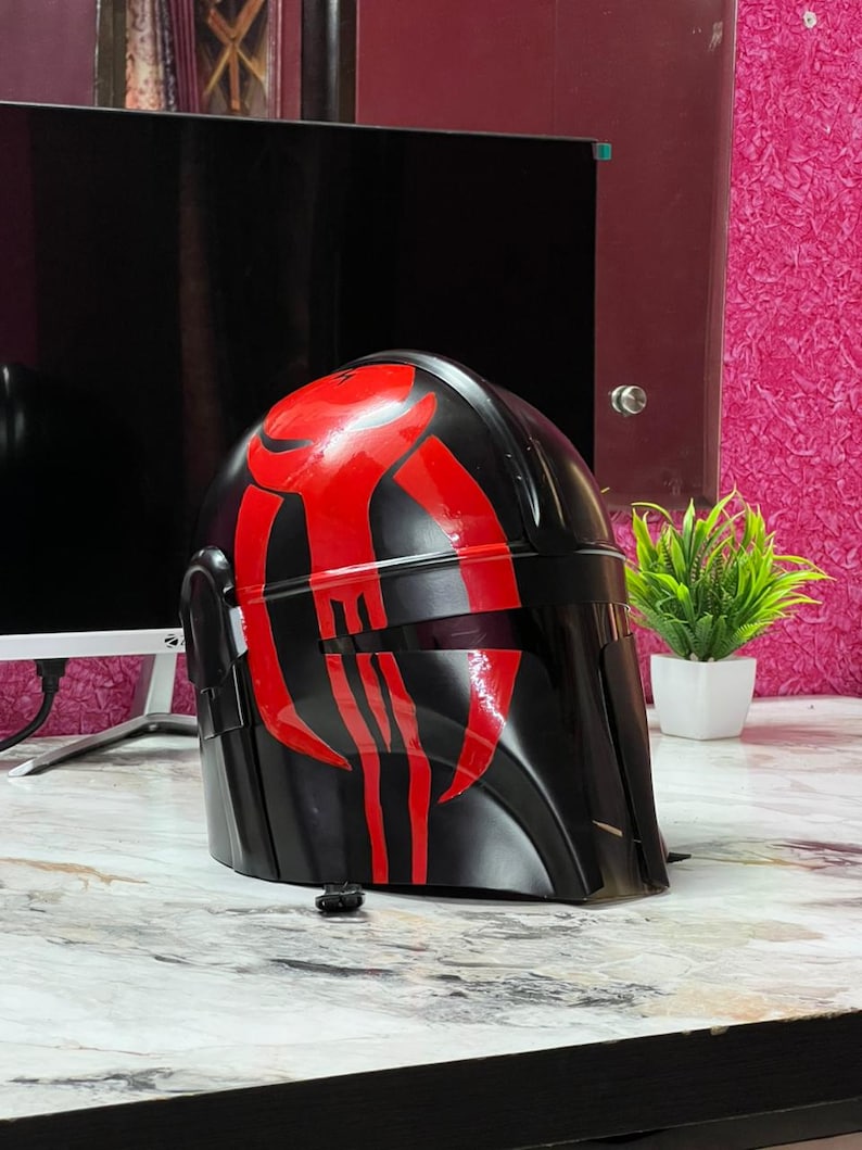 May include: A black helmet with red markings, likely a costume piece. The helmet has a smooth, glossy finish and a visor. The red design is a stylized pattern on the front and top of the helmet. The helmet is sitting on a marble-patterned surface.