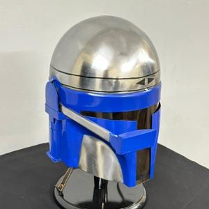 Op de afbeelding: Een zilveren en blauwe Mandalorian helm. De helm heeft een gepolijste zilveren koepel en een blauw vizier en frontplaat. De helm wordt op een zwarte standaard tentoongesteld.