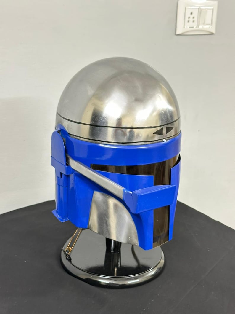 Op de afbeelding: Een zilveren en blauwe Mandalorian helm. De helm heeft een gepolijste zilveren koepel en een blauw vizier en frontplaat. De helm wordt op een zwarte standaard tentoongesteld.