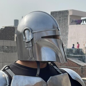 May include: A silver-colored Mandalorian helmet with a T-shaped visor and ear pieces. The helmet is part of a full set of armor, including chest and shoulder plates. The armor is worn over a black shirt.