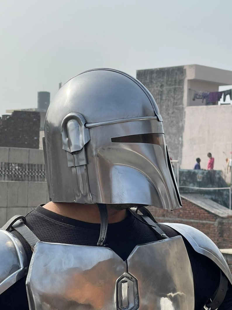 May include: A silver-colored Mandalorian helmet with a T-shaped visor and ear pieces. The helmet is part of a full set of armor, including chest and shoulder plates. The armor is worn over a black shirt.