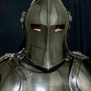 May include: A close-up of a black medieval knight's helmet and armor. The helmet has a cross design and eye slits. The armor is made of metal and has rivets and straps. The background is black.