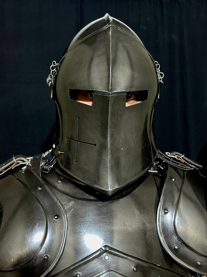 May include: A close-up of a black medieval knight's helmet and armor. The helmet has a cross design and eye slits. The armor is made of metal and has rivets and straps. The background is black.
