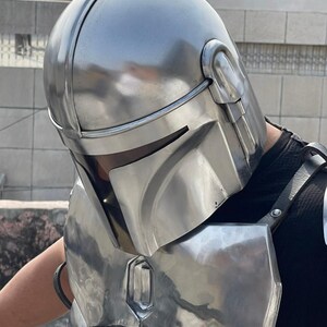 May include: A silver-colored Mandalorian helmet and chest armor set. The helmet features a rounded top with a central ridge and visor. The chest armor has a rectangular shape with a metallic sheen, and is secured with black straps.