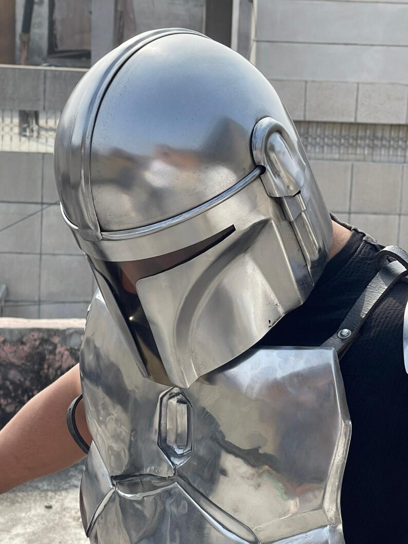 May include: A silver-colored Mandalorian helmet and chest armor set. The helmet features a rounded top with a central ridge and visor. The chest armor has a rectangular shape with a metallic sheen, and is secured with black straps.