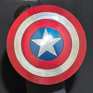 May include: A Captain America shield featuring a red, white, and blue color scheme. The shield has a circular design with concentric rings of red, silver, and blue, and a white star in the center. The shield is a prop or collectible.