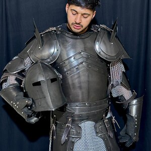 May include: A full suit of black medieval armor, including a helmet, chest plate, shoulder guards, and arm and leg protection. The armor is made of metal and chainmail, with leather straps and rivets. The helmet has a visor with eye slits.