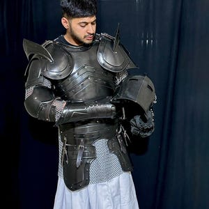 May include: A dark gray medieval knight's armor set, including a breastplate, shoulder guards, and helmet. The armor is detailed with rivets and straps, and is paired with chainmail and a white skirt. The knight is holding the helmet.