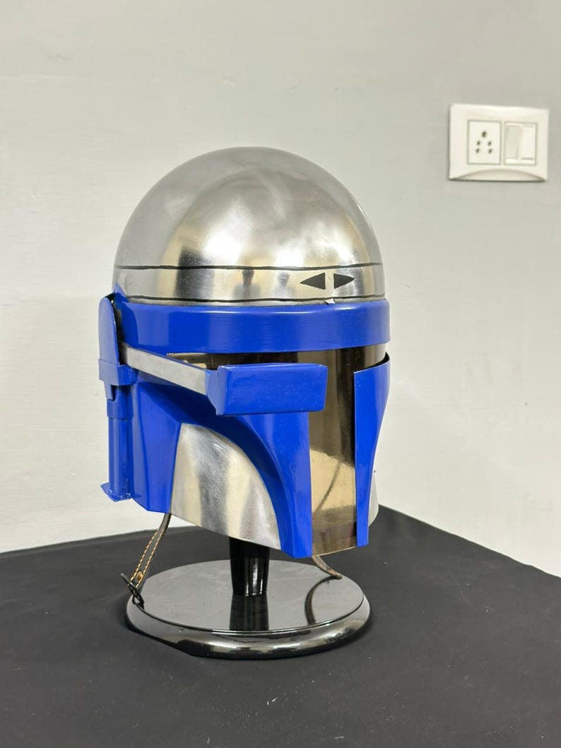 Op de afbeelding: Een zilveren en blauwe helm, waarschijnlijk een rekwisiet of verzamelobject. De helm heeft een afgeronde bovenkant en blauwe vizier- en wangstukken. Hij wordt gepresenteerd op een zwarte standaard, wat suggereert dat het een decoratief item of onderdeel van een kostuum is.