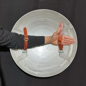 May include: A silver-colored, circular shield with concentric ridges. It features two brown leather straps with gold-colored buckles for securing the shield. The shield is held against a black background.