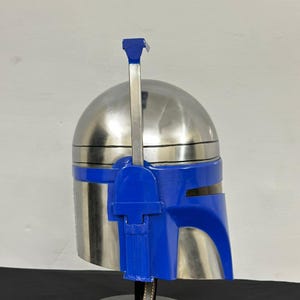 Op de afbeelding: Een zilverkleurige helm met blauwe accenten, inclusief vizier en een verticale antenne. De helm wordt gepresenteerd op een zwarte standaard met een leren riem.