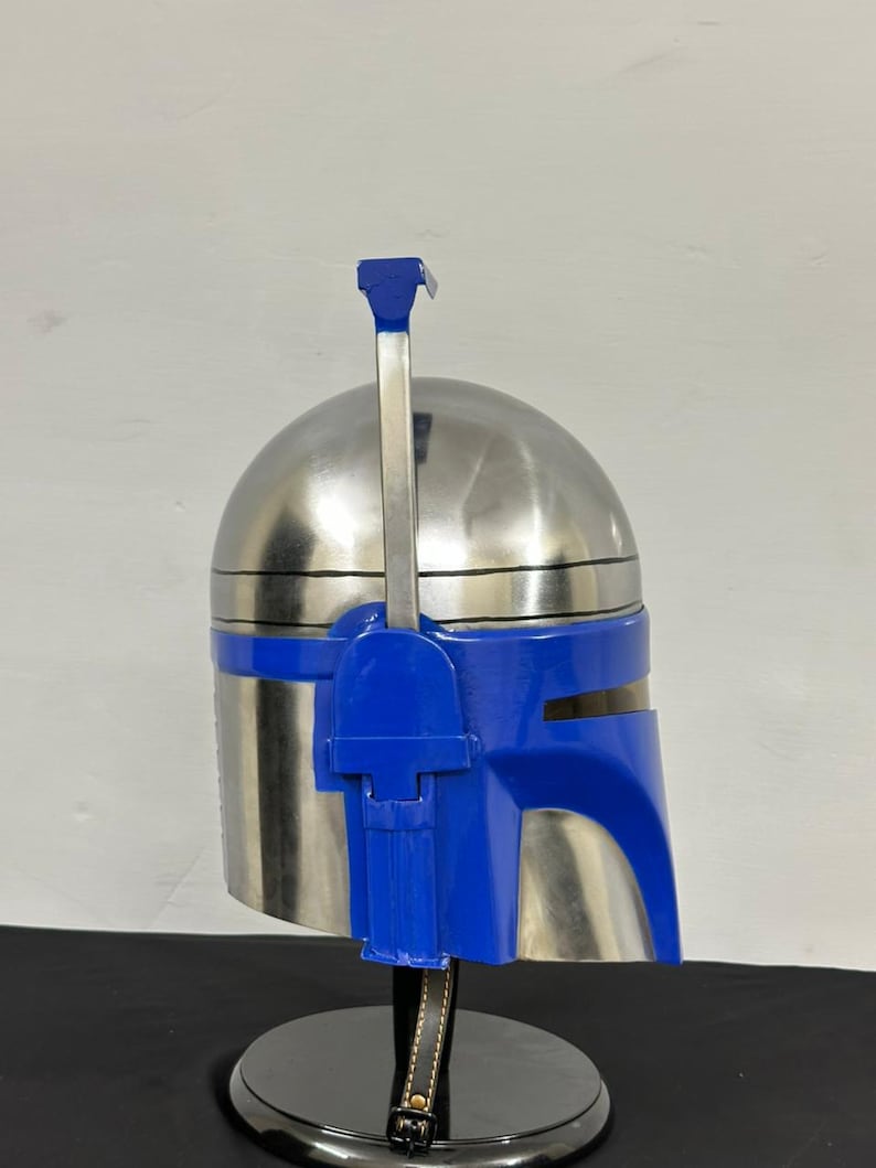 Op de afbeelding: Een zilverkleurige helm met blauwe accenten, inclusief vizier en een verticale antenne. De helm wordt gepresenteerd op een zwarte standaard met een leren riem.