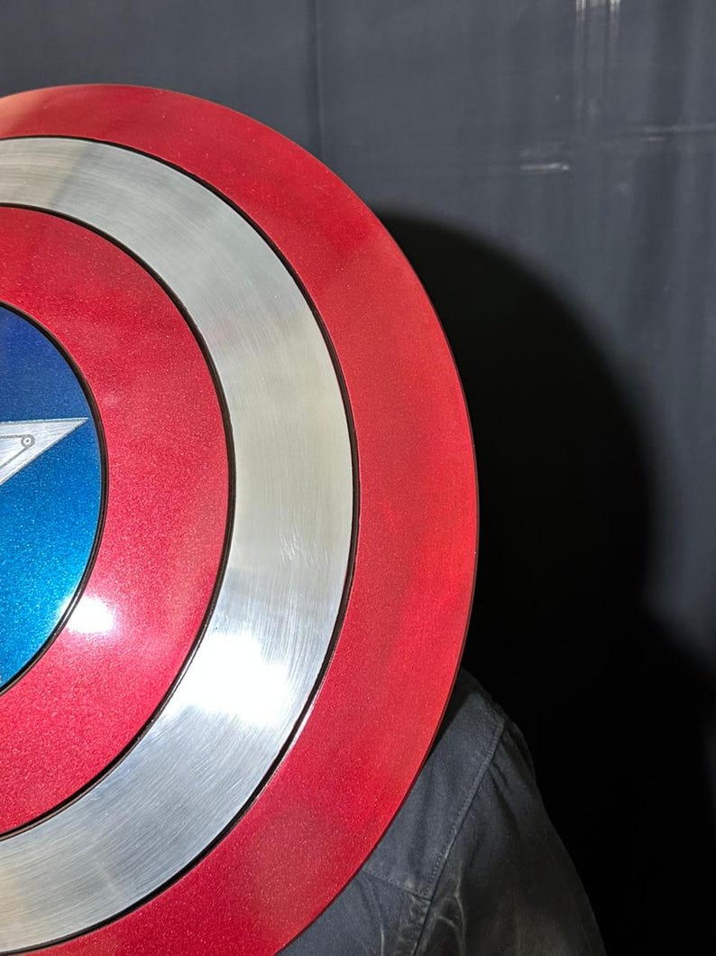 May include: A red, white, and blue shield with a star emblem. The shield has concentric circles of red, silver, and red, with a blue center featuring a white star. The shield is partially obscuring a person wearing a black shirt.