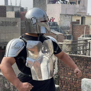 May include: A silver-colored Mandalorian helmet and armor set. The helmet has a distinctive visor and ear pieces. The chest plate and shoulder pads are also silver, with a metallic sheen. The armor is worn over a black shirt.