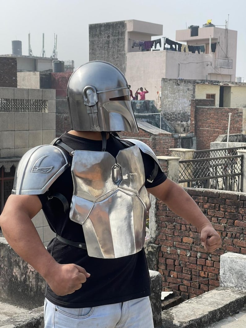 May include: A silver-colored Mandalorian helmet and armor set. The helmet has a distinctive visor and ear pieces. The chest plate and shoulder pads are also silver, with a metallic sheen. The armor is worn over a black shirt.