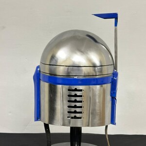 Op de afbeelding: Een zilveren en blauwe helm, ontworpen in de stijl van een Mandalorian helm. De helm heeft een afgeronde bovenkant, een blauwe band en blauwe accenten aan de zijkanten. Hij staat op een zwarte standaard.