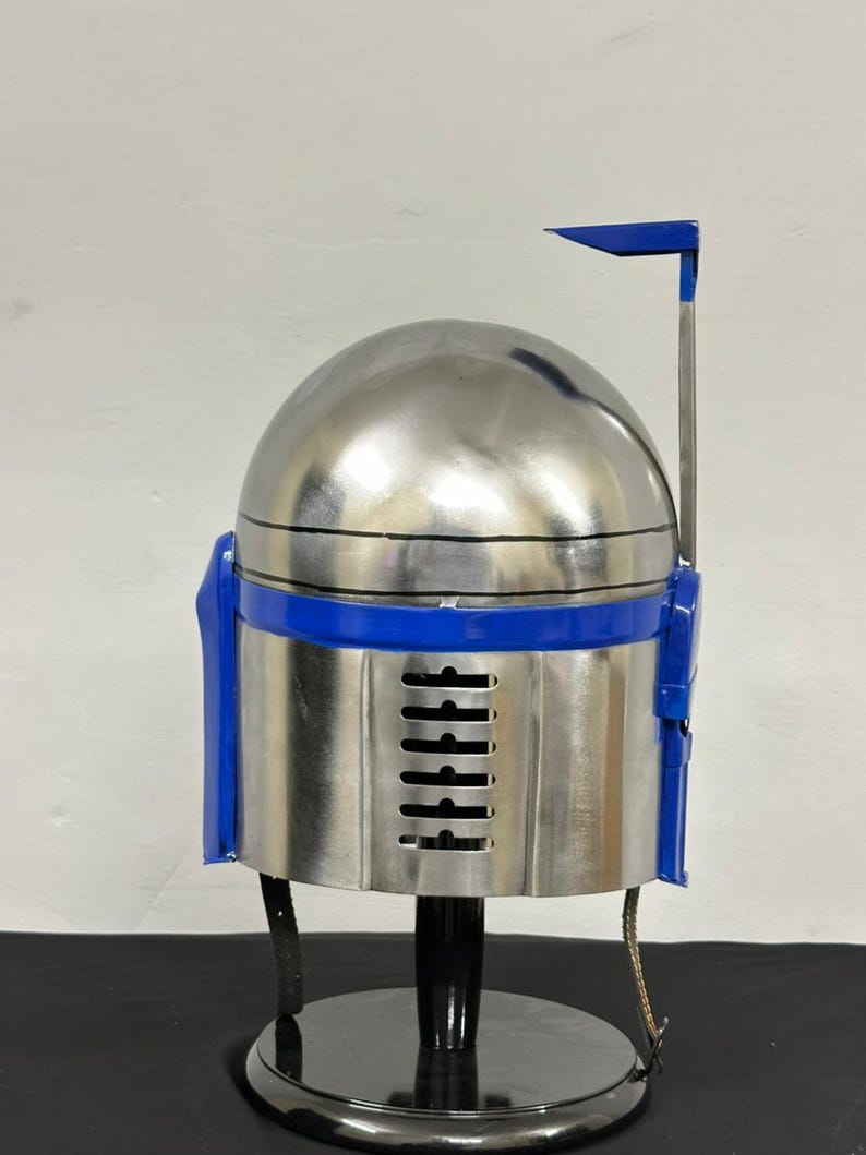 Op de afbeelding: Een zilveren en blauwe helm, ontworpen in de stijl van een Mandalorian helm. De helm heeft een afgeronde bovenkant, een blauwe band en blauwe accenten aan de zijkanten. Hij staat op een zwarte standaard.