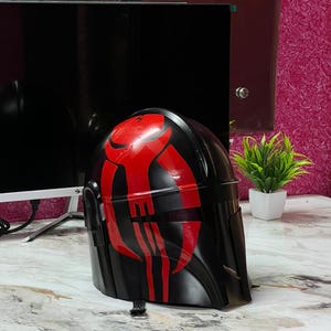 May include: A black helmet with a red graphic design. The helmet is glossy and appears to be made of plastic. The red design is a series of vertical and horizontal lines. The helmet is sitting on a marble-patterned surface.