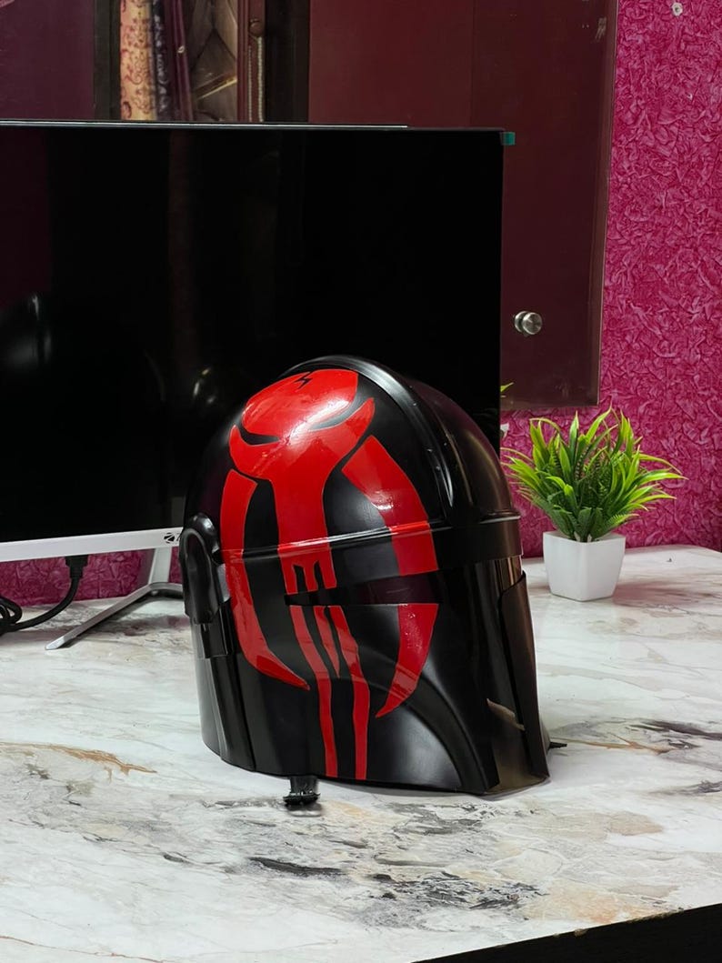 May include: A black helmet with a red graphic design. The helmet is glossy and appears to be made of plastic. The red design is a series of vertical and horizontal lines. The helmet is sitting on a marble-patterned surface.