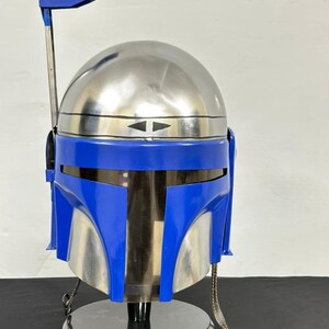 Op de afbeelding: Een zilveren en blauwe helm, die doet denken aan een Mandalorian helm, staat op een zwarte cirkelvormige basis. De helm heeft een blauw vizier en een zilveren koepel. Een zilveren antenne strekt zich uit vanaf de bovenkant.