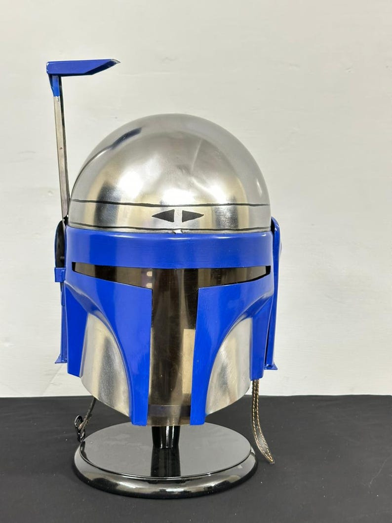 Op de afbeelding: Een zilveren en blauwe helm, die doet denken aan een Mandalorian helm, staat op een zwarte cirkelvormige basis. De helm heeft een blauw vizier en een zilveren koepel. Een zilveren antenne strekt zich uit vanaf de bovenkant.