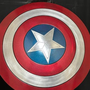 May include: A red, white, and blue Captain America shield. The shield has a red outer ring, a silver middle ring, and a blue center with a white star. The shield is a circular shape.