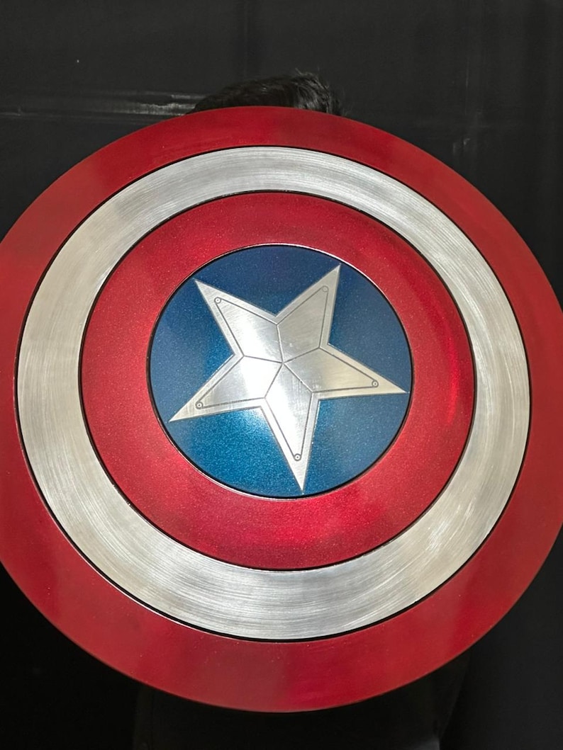 May include: A red, white, and blue Captain America shield. The shield has a red outer ring, a silver middle ring, and a blue center with a white star. The shield is a circular shape.