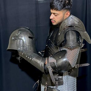 May include: Black medieval armor set, including a helmet, shoulder guards, arm guards, and chest plate. The armor features leather straps, metal rivets, and chainmail accents. The helmet has a visor and is held in the hand.