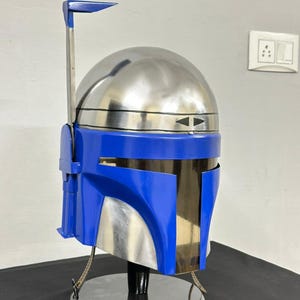 Op de afbeelding: Een blauw en zilverkleurige helm met een kenmerkend ontwerp, met een afgeronde bovenkant en een hoekig vizier. De helm is gemonteerd op een zwarte standaard, die de metallic en glanzende afwerking benadrukt. De helm heeft een zilveren antenne en een blauw accent.